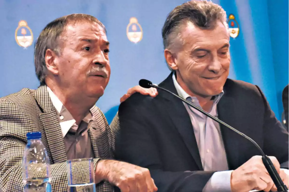 Two men in suits sitting together at a press conference with a blue background and official emblems.