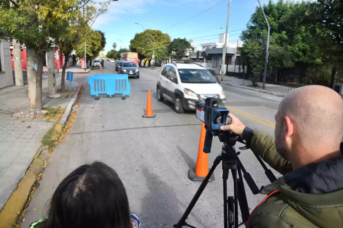 A man uses a speed radar on a street where there are orange cones and a blue barrier as cars pass by. A man uses a speed radar on a street where there are orange cones and a blue barrier as cars pass by.