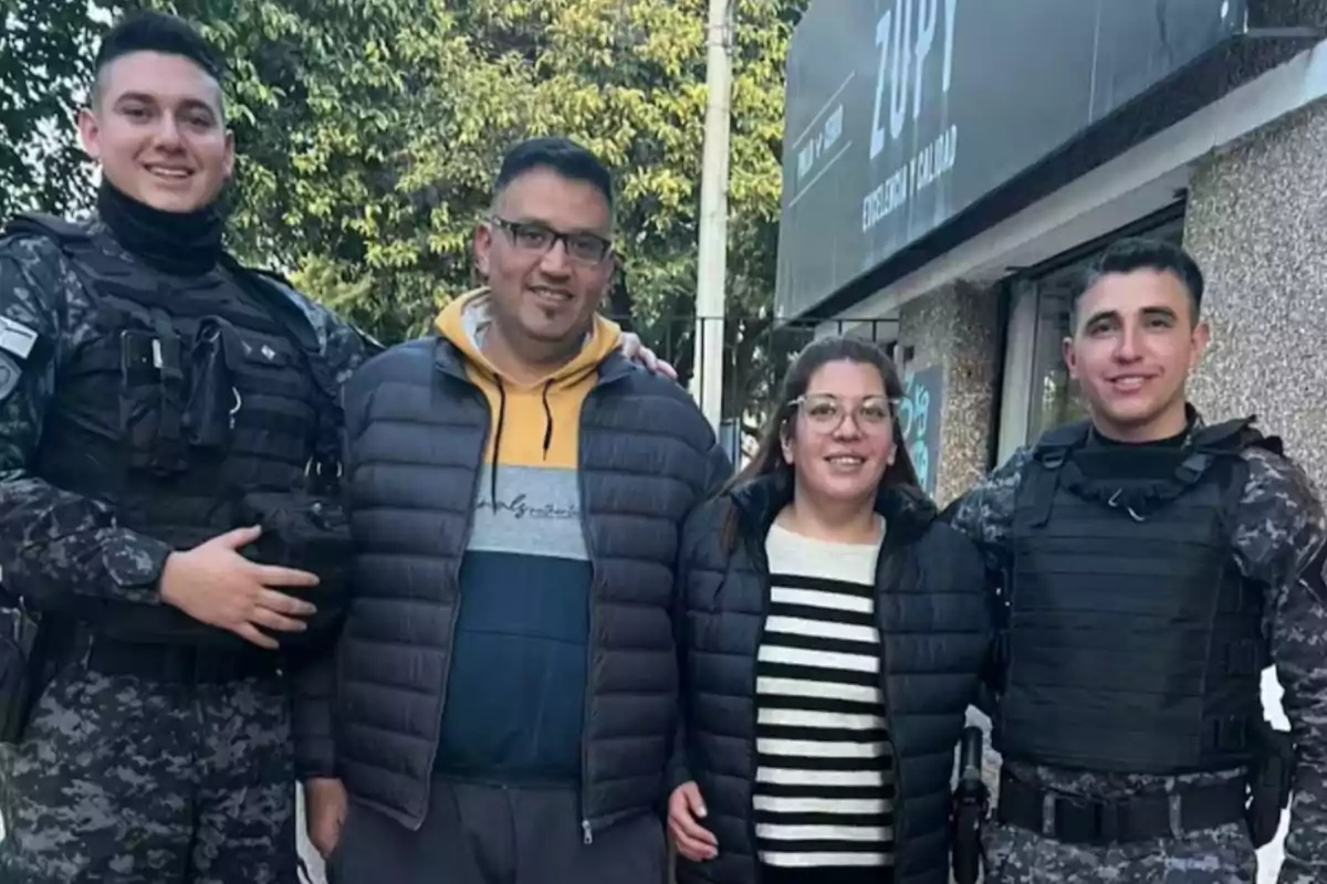Four people are smiling and posing on the street; two of them are wearing police uniforms and the other two are dressed in casual clothes with warm jackets. In the background, there is a store sign and some trees.