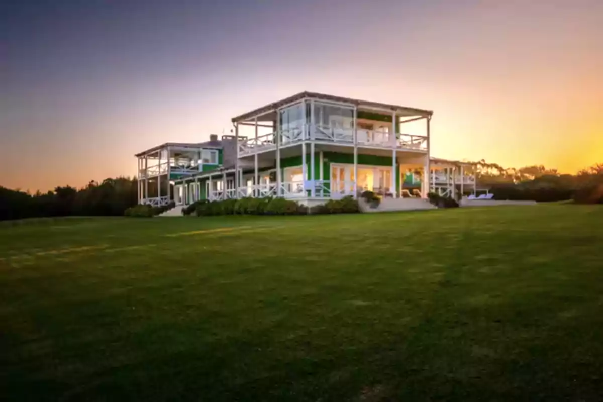 Modern two-story house with large windows and a terrace, surrounded by grass and greenery at sunset Modern two-story house with large windows and a terrace, surrounded by grass and greenery at sunset