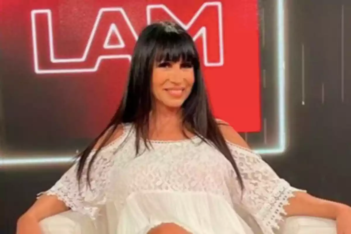Long-haired, dark-haired woman with bangs, wearing a white lace blouse, sitting and smiling on a television set with a red background and white letters. Long-haired, dark-haired woman with bangs, wearing a white lace blouse, sitting and smiling on a television set with a red background and white letters.