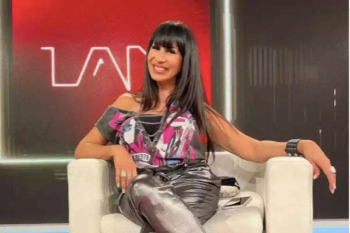 Woman with long dark hair sitting on a white armchair, smiling, with a red background and blurred white letters Woman with long dark hair sitting on a white armchair, smiling, with a red background and blurred white letters