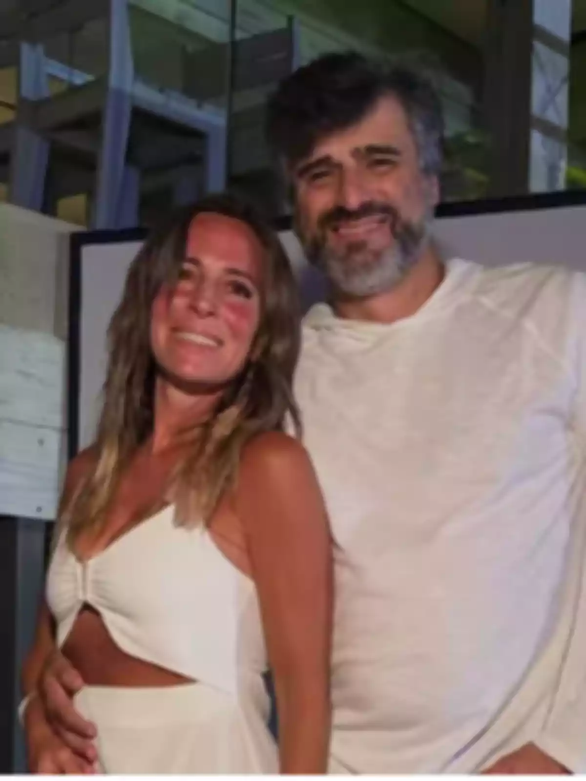 A couple smiles while hugging as they pose for the photo, both dressed in white in a modern indoor setting. A couple smiles while hugging as they pose for the photo, both dressed in white in a modern indoor setting.
