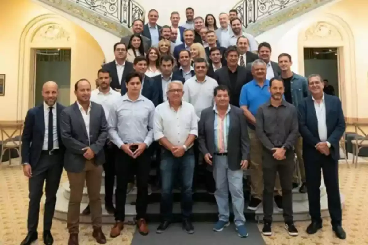 Un grupo numeroso de personas posando para una foto en el interior de un edificio elegante con escaleras y barandales decorativos al fondo