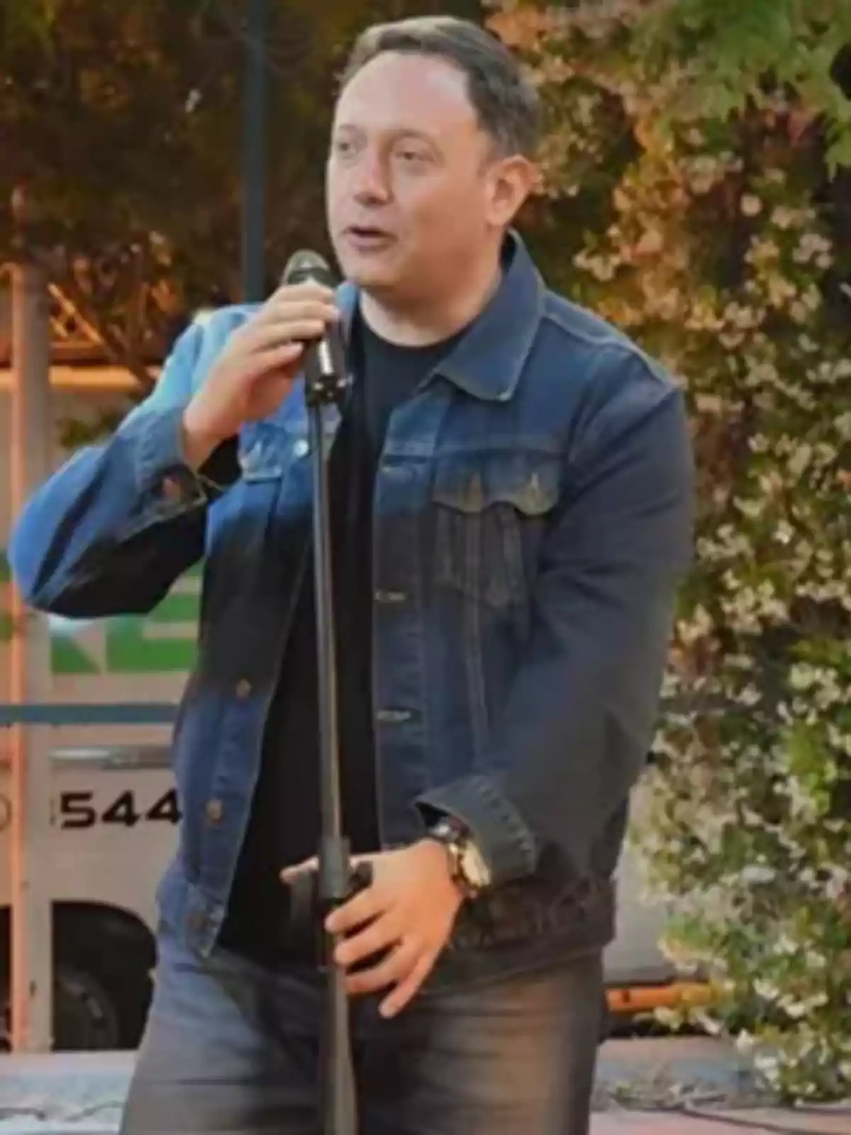 Hombre de cabello corto usando chaqueta de mezclilla y sosteniendo un micrófono mientras habla al aire libre Hombre de cabello corto usando chaqueta de mezclilla y sosteniendo un micrófono mientras habla al aire libre