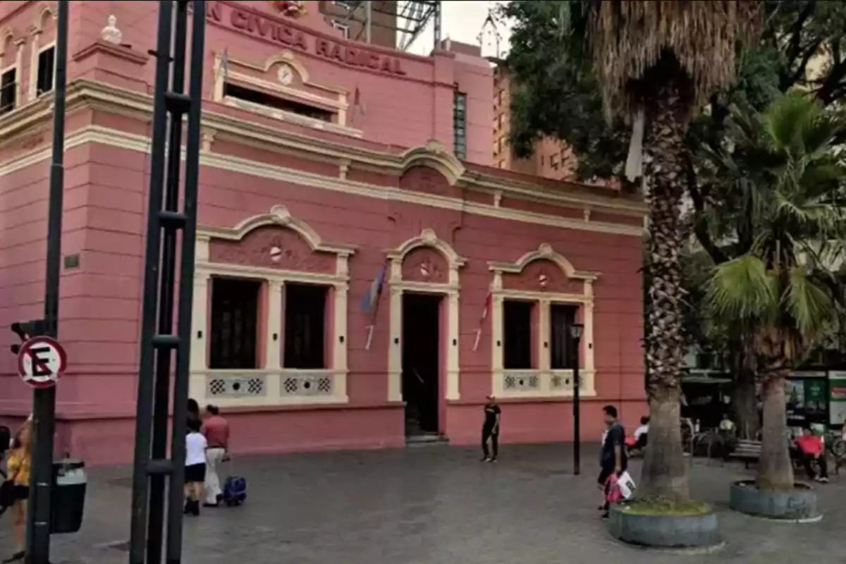 Edificio antiguo de color rosa con detalles en blanco ubicado en una esquina de una plaza con palmeras y personas caminando y sentadas alrededor Edificio antiguo de color rosa con detalles en blanco ubicado en una esquina de una plaza con palmeras y personas caminando y sentadas alrededor