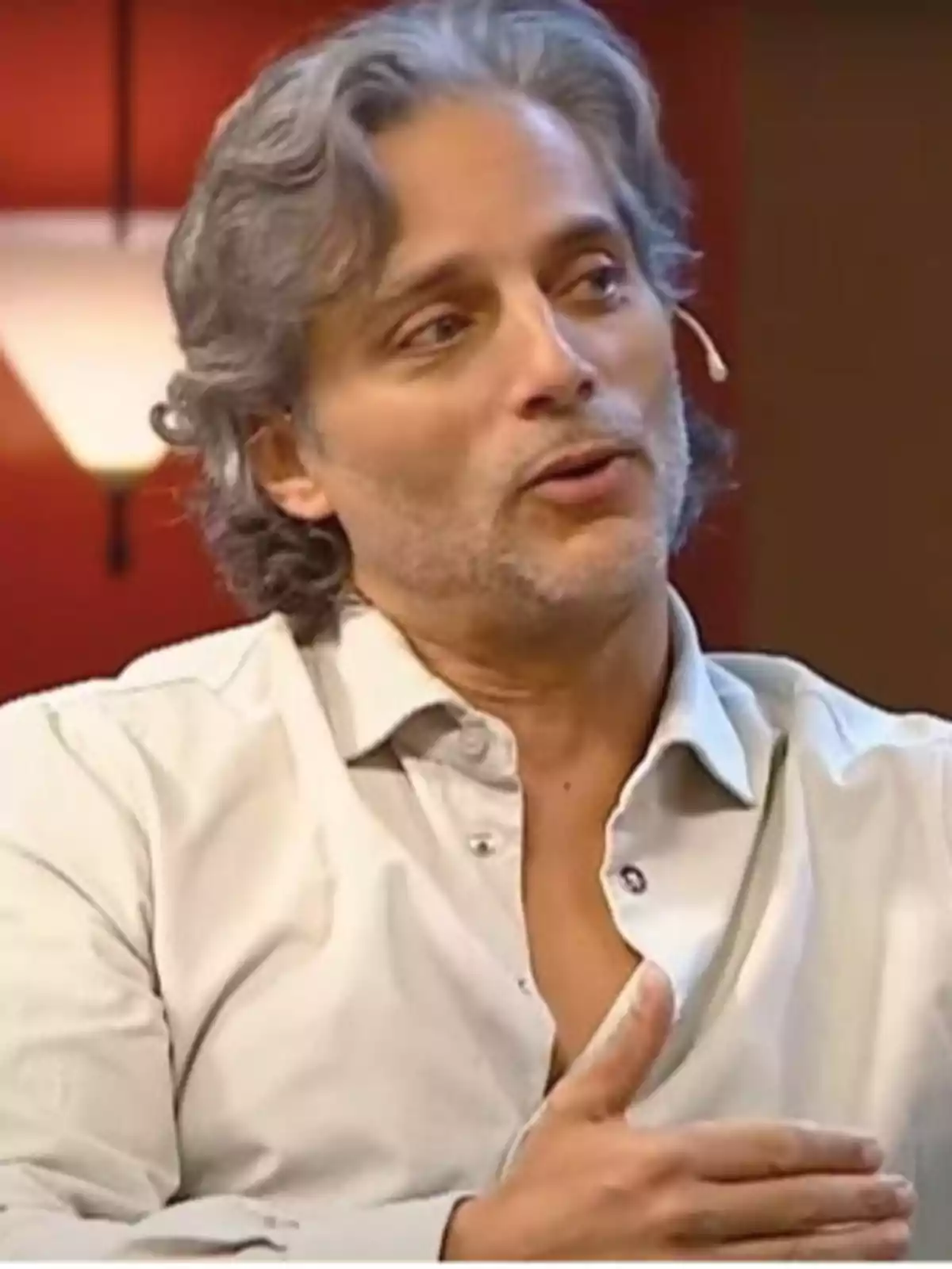 Gray-haired, wavy-haired man wearing a light-colored jersey and a headset microphone while speaking on a television set with a red background. Gray-haired, wavy-haired man wearing a light-colored jersey and a headset microphone while speaking on a television set with a red background.