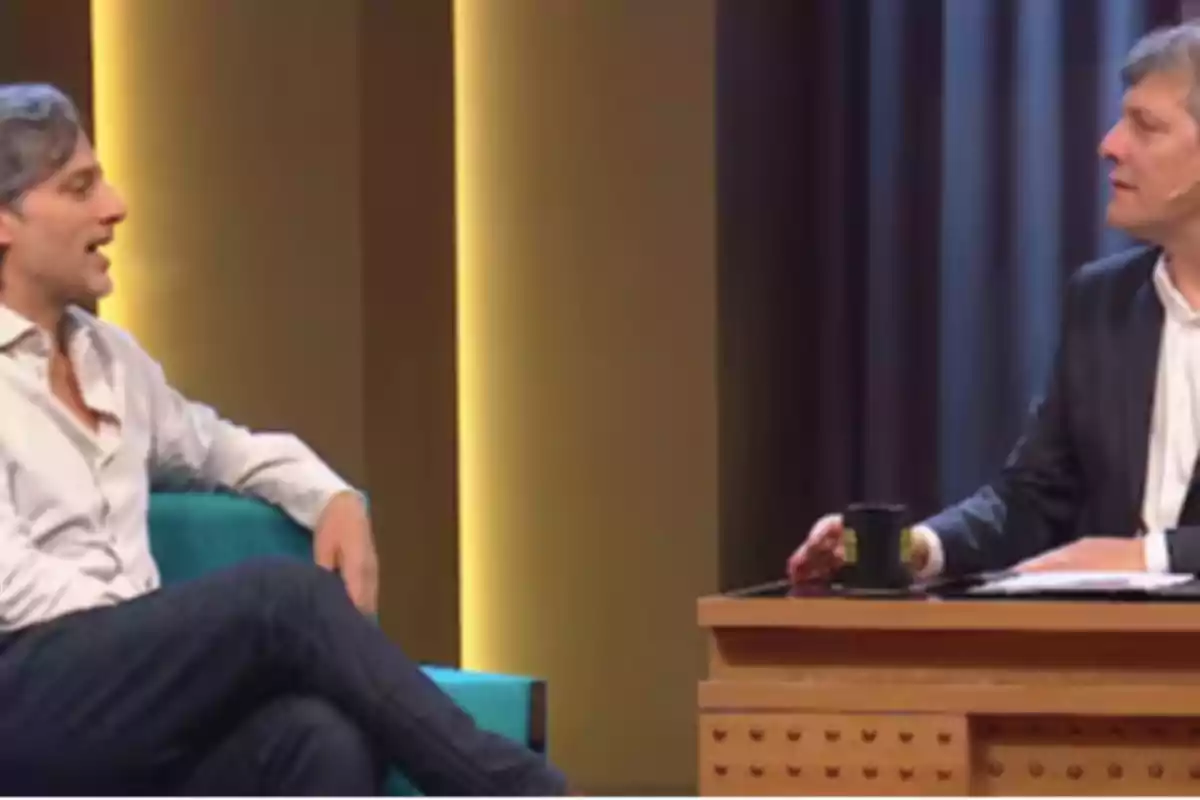 Two men are talking on a talk show set, one sitting in an armchair and the other behind a desk with a cup and some papers. Two men are talking on a talk show set, one sitting in an armchair and the other behind a desk with a cup and some papers.