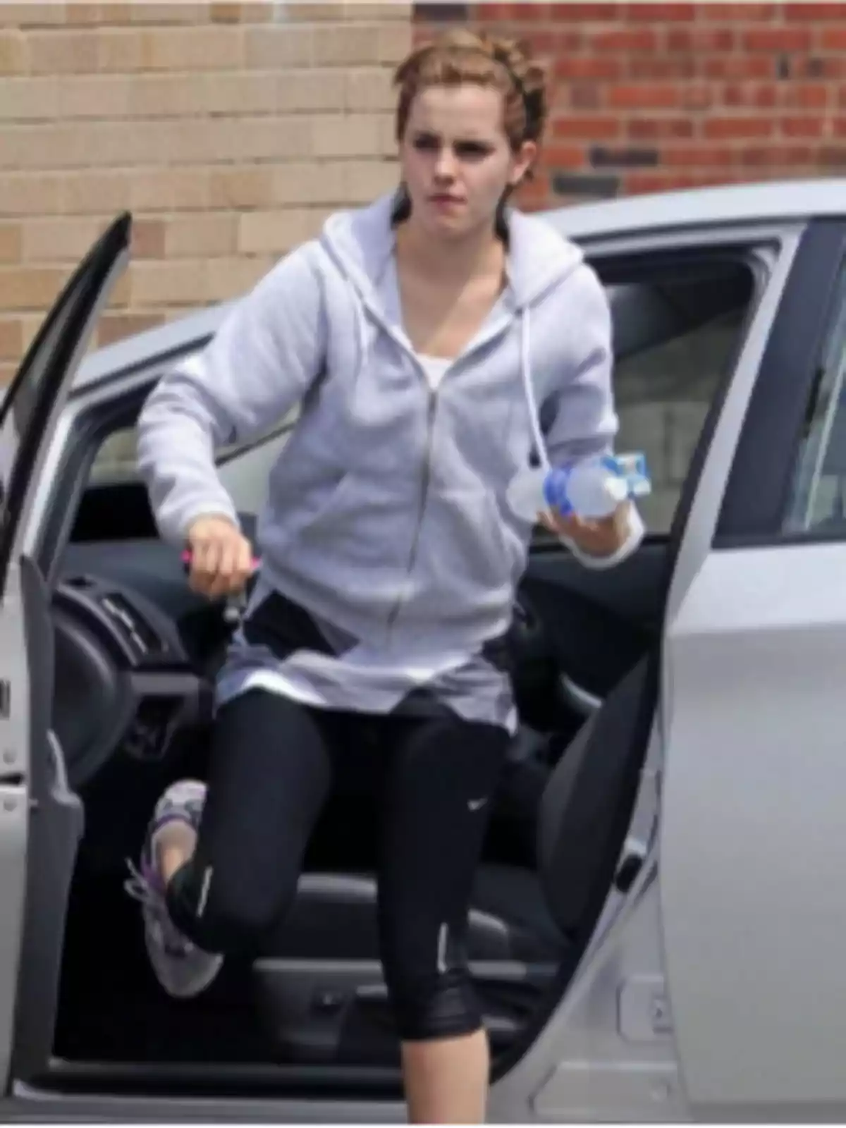 Mujer joven con ropa deportiva saliendo de un auto plateado mientras sostiene una botella de agua Mujer joven con ropa deportiva saliendo de un auto plateado mientras sostiene una botella de agua