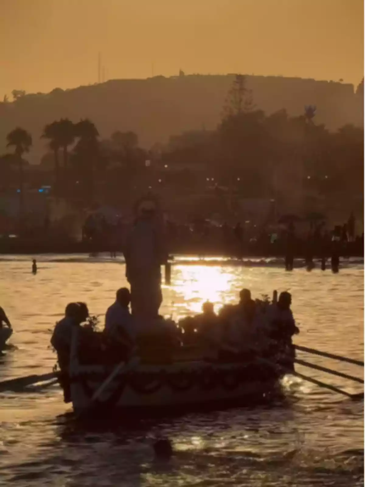Boat with several people rowing at sunset on the sea, with a religious figure in the center and hills in the background Boat with several people rowing at sunset on the sea, with a religious figure in the center and hills in the background