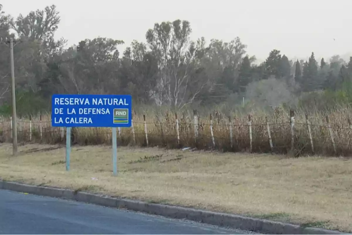 Blue sign indicating the entrance to the La Calera Defense Nature Reserve next to a road and a wire fence with vegetation in the background Blue sign indicating the entrance to the La Calera Defense Nature Reserve next to a road and a wire fence with vegetation in the background