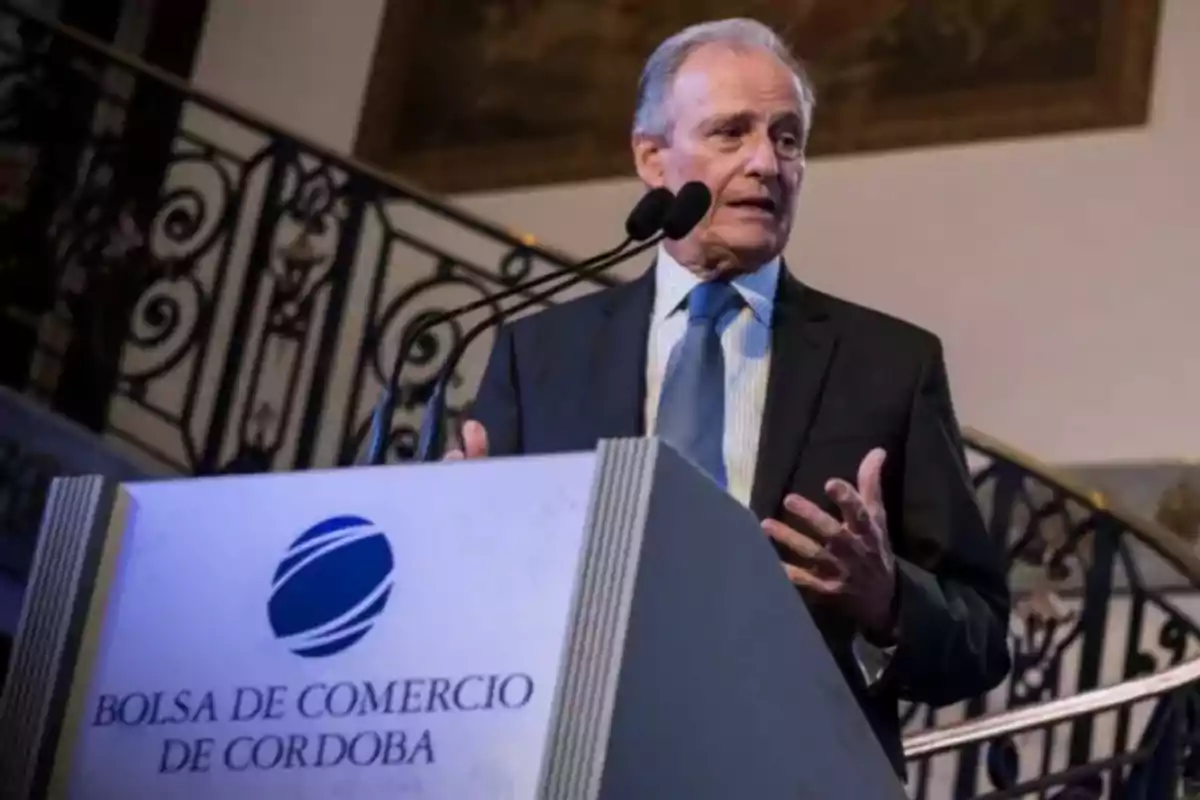 Older man in a suit speaking at a podium of the Córdoba Stock Exchange with a staircase in the background Older man in a suit speaking at a podium of the Córdoba Stock Exchange with a staircase in the background