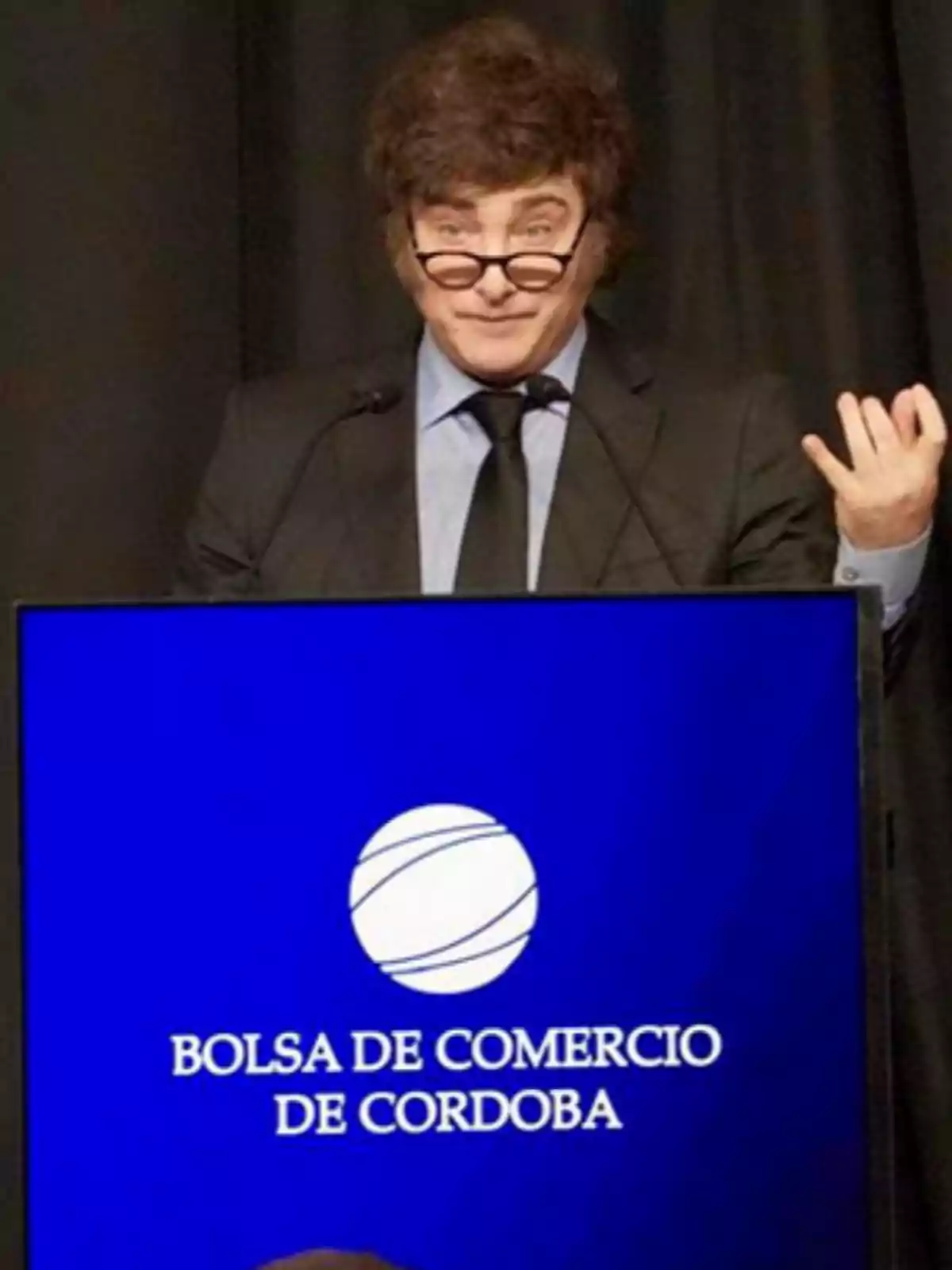 Person with glasses speaking at a blue podium that has the text Córdoba Stock Exchange Person with glasses speaking at a blue podium that has the text Córdoba Stock Exchange