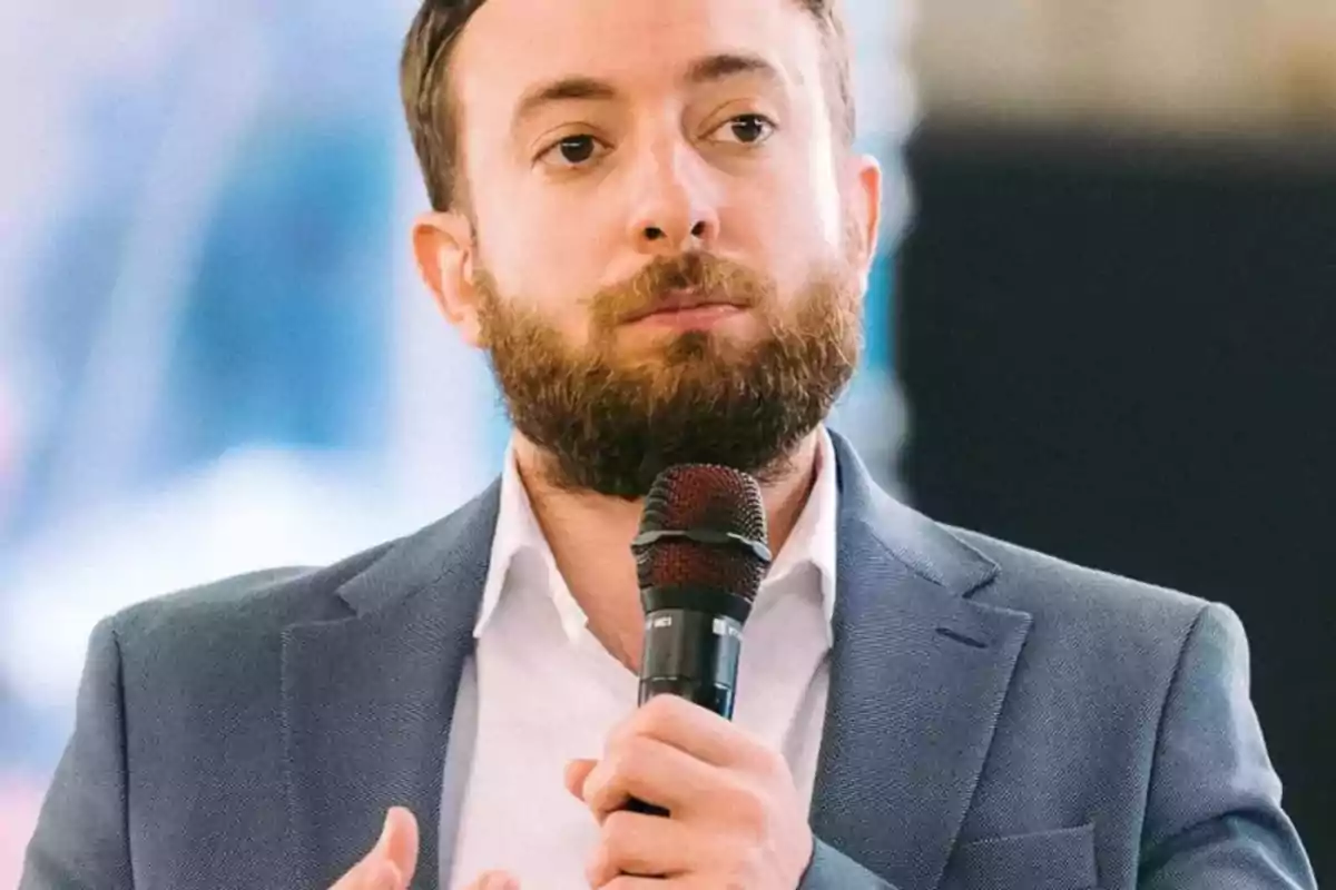 Hombre con barba y saco azul hablando mientras sostiene un micrófono Hombre con barba y saco azul hablando mientras sostiene un micrófono