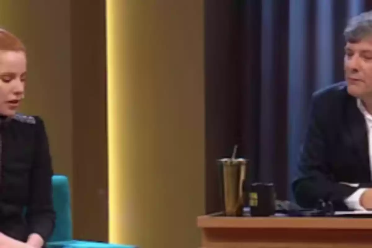 Two people sitting on a television set during an interview, one of them is in a blue armchair and the other is behind a desk with a cup and a metal tumbler. Two people sitting on a television set during an interview, one of them is in a blue armchair and the other is behind a desk with a cup and a metal tumbler.