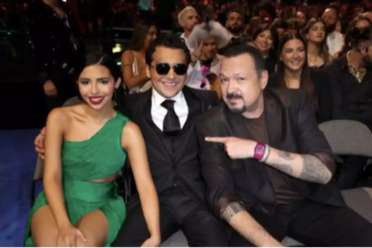 Tres personas sentadas en primera fila en un evento formal, una mujer con vestido verde, un hombre con traje y gafas oscuras y otro hombre con chaqueta oscura señalando al del centro, rodeados de más asistentes en el fondo. Tres personas sentadas en primera fila en un evento formal, una mujer con vestido verde, un hombre con traje y gafas oscuras y otro hombre con chaqueta oscura señalando al del centro, rodeados de más asistentes en el fondo.
