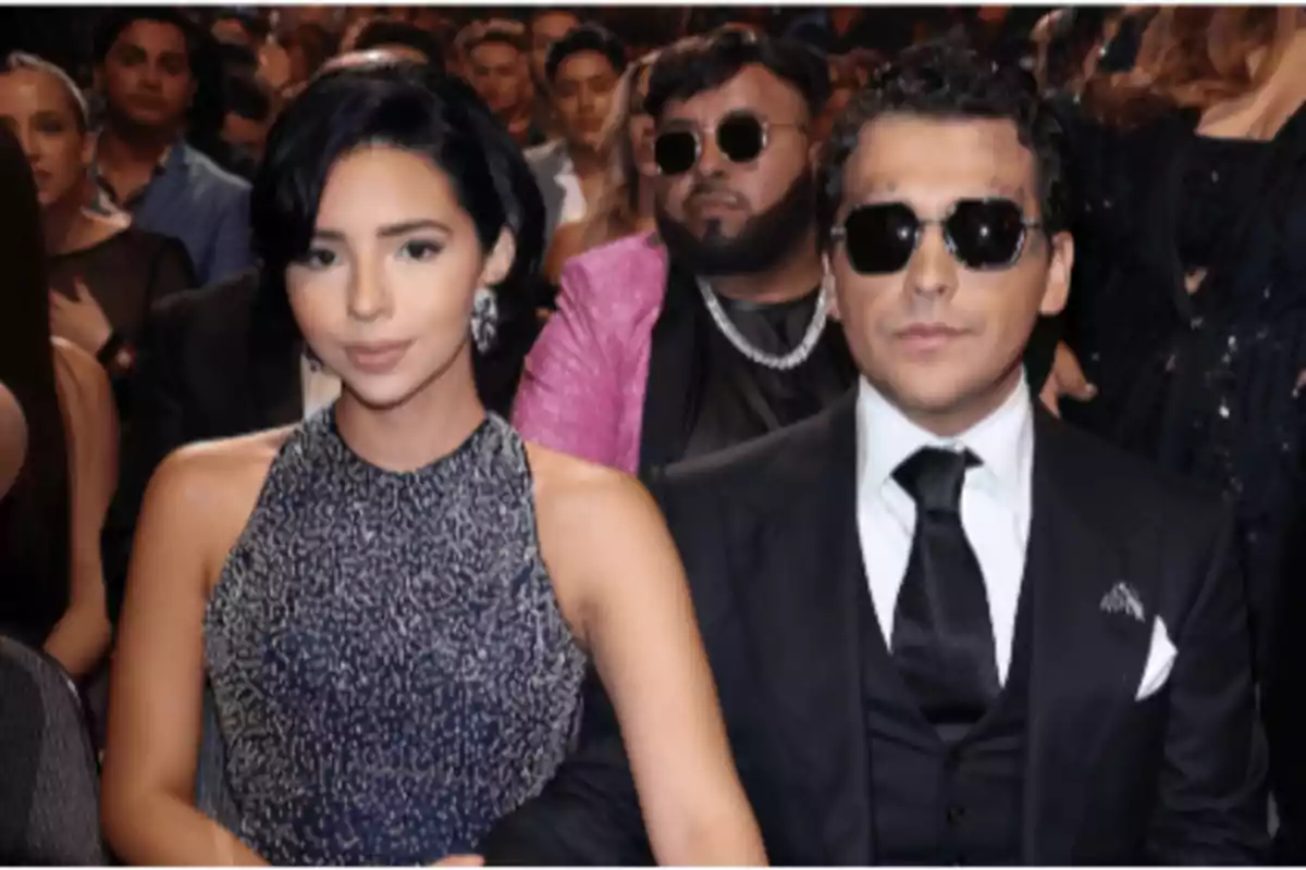 Una pareja elegantemente vestida está sentada en un evento rodeada de otras personas, el hombre lleva gafas oscuras y traje negro, la mujer lleva un vestido azul brillante
