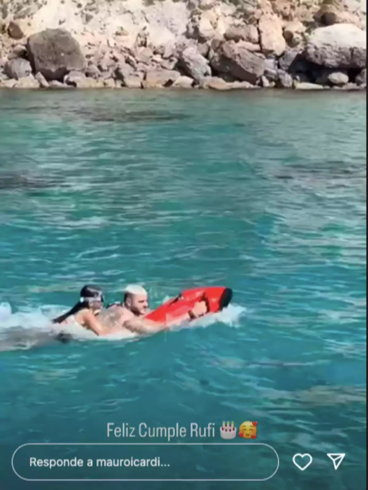 Dos personas nadan en el mar cerca de unas rocas, una de ellas sostiene una tabla de rescate roja Dos personas nadan en el mar cerca de unas rocas, una de ellas sostiene una tabla de rescate roja
