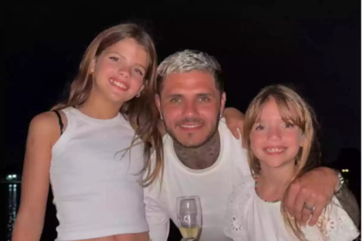 Tres personas sonríen abrazadas frente a un fondo oscuro, dos niñas y un hombre con una copa en la mano, todos vestidos de blanco. Tres personas sonríen abrazadas frente a un fondo oscuro, dos niñas y un hombre con una copa en la mano, todos vestidos de blanco.