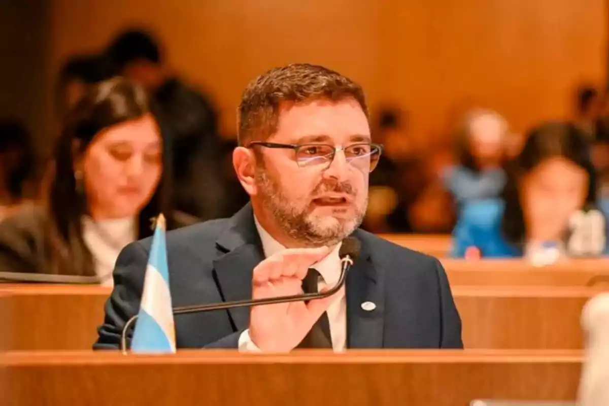 Hombre con gafas y barba hablando en un micrófono en una sala de conferencias con una bandera argentina sobre la mesa Hombre con gafas y barba hablando en un micrófono en una sala de conferencias con una bandera argentina sobre la mesa