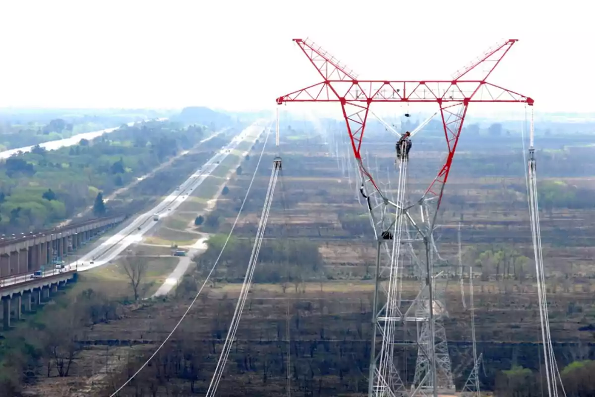 Torre de alta tensión con trabajadores realizando mantenimiento mientras al fondo se observa una carretera y un paisaje rural Torre de alta tensión con trabajadores realizando mantenimiento mientras al fondo se observa una carretera y un paisaje rural