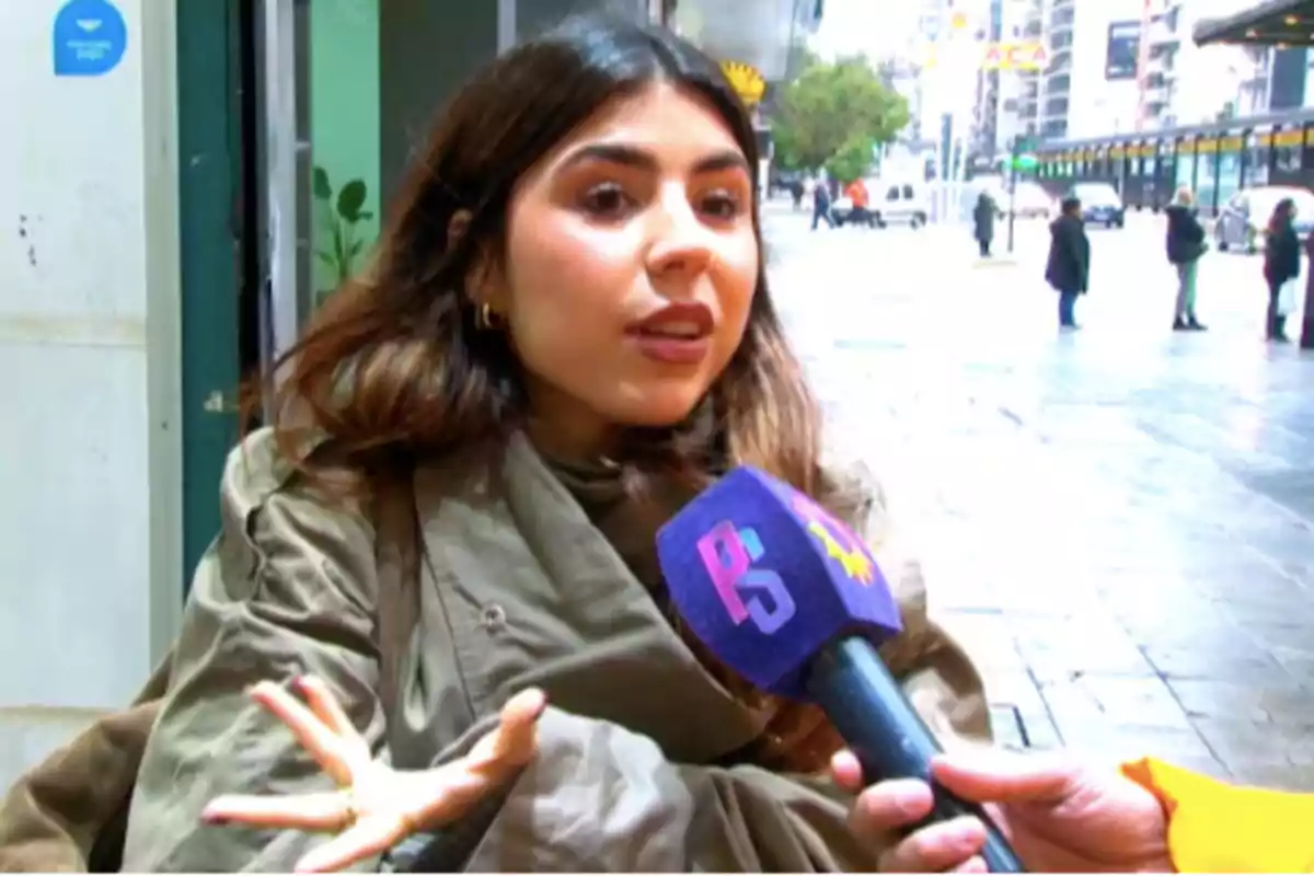 Mujer joven con cabello largo y suelto siendo entrevistada en la calle mientras sostiene un micrófono azul con letras de colores Mujer joven con cabello largo y suelto siendo entrevistada en la calle mientras sostiene un micrófono azul con letras de colores