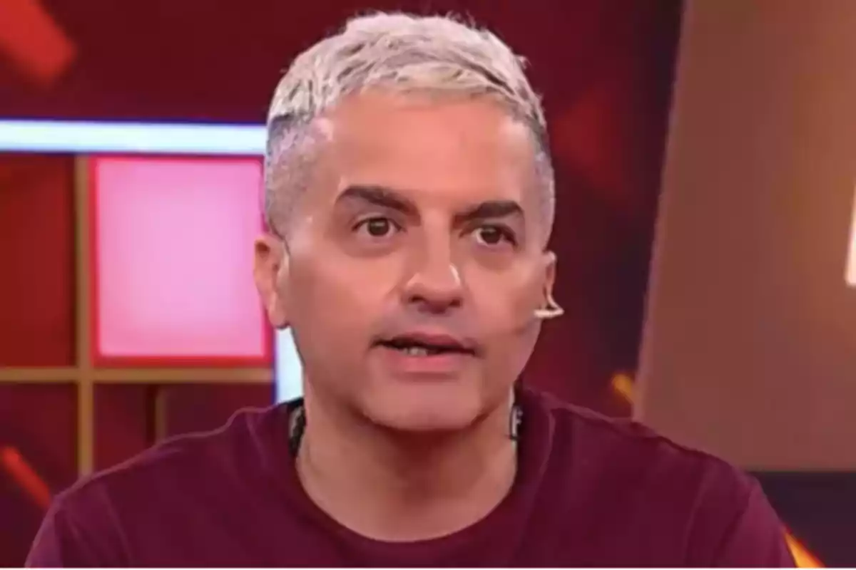 Short-haired, platinum blonde man speaking on a television set with a red background and bright lights