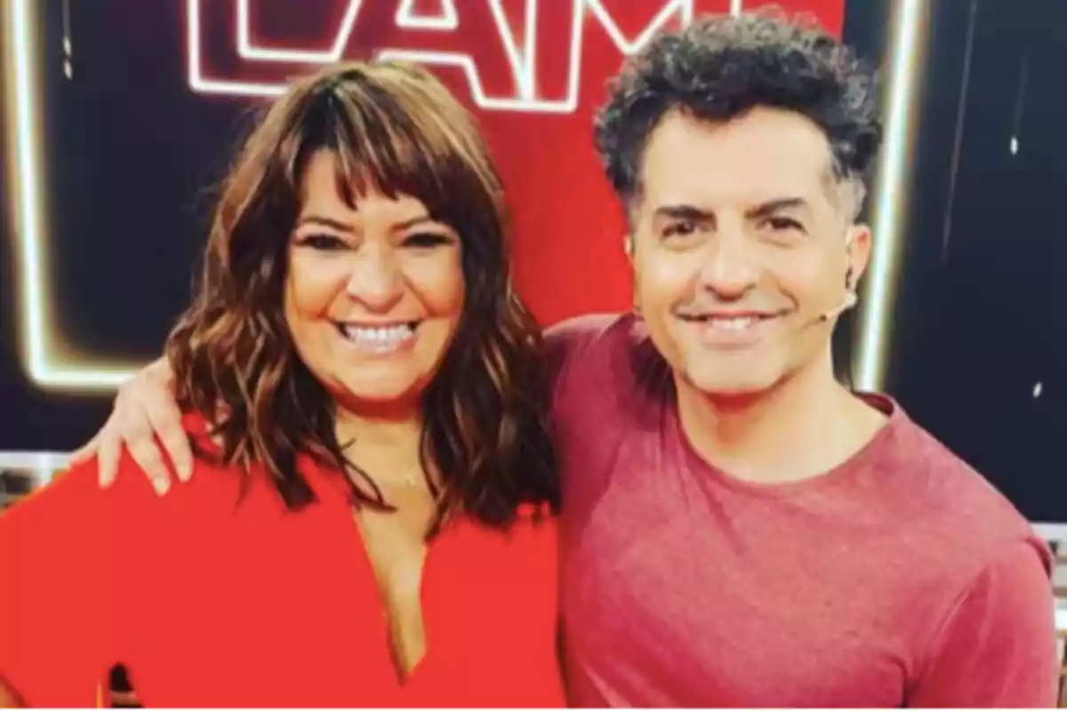 Dos personas sonrientes posan abrazadas en un set de televisión con fondo rojo y letras luminosas. Dos personas sonrientes posan abrazadas en un set de televisión con fondo rojo y letras luminosas.