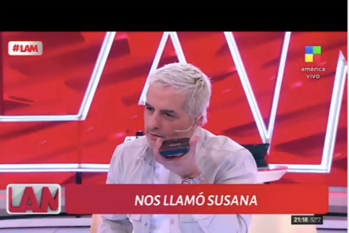 Conductor de televisión de cabello canoso sostiene un teléfono móvil durante una llamada en el programa LAM con un fondo rojo y un cartel que dice nos llamó Susana Conductor de televisión de cabello canoso sostiene un teléfono móvil durante una llamada en el programa LAM con un fondo rojo y un cartel que dice nos llamó Susana