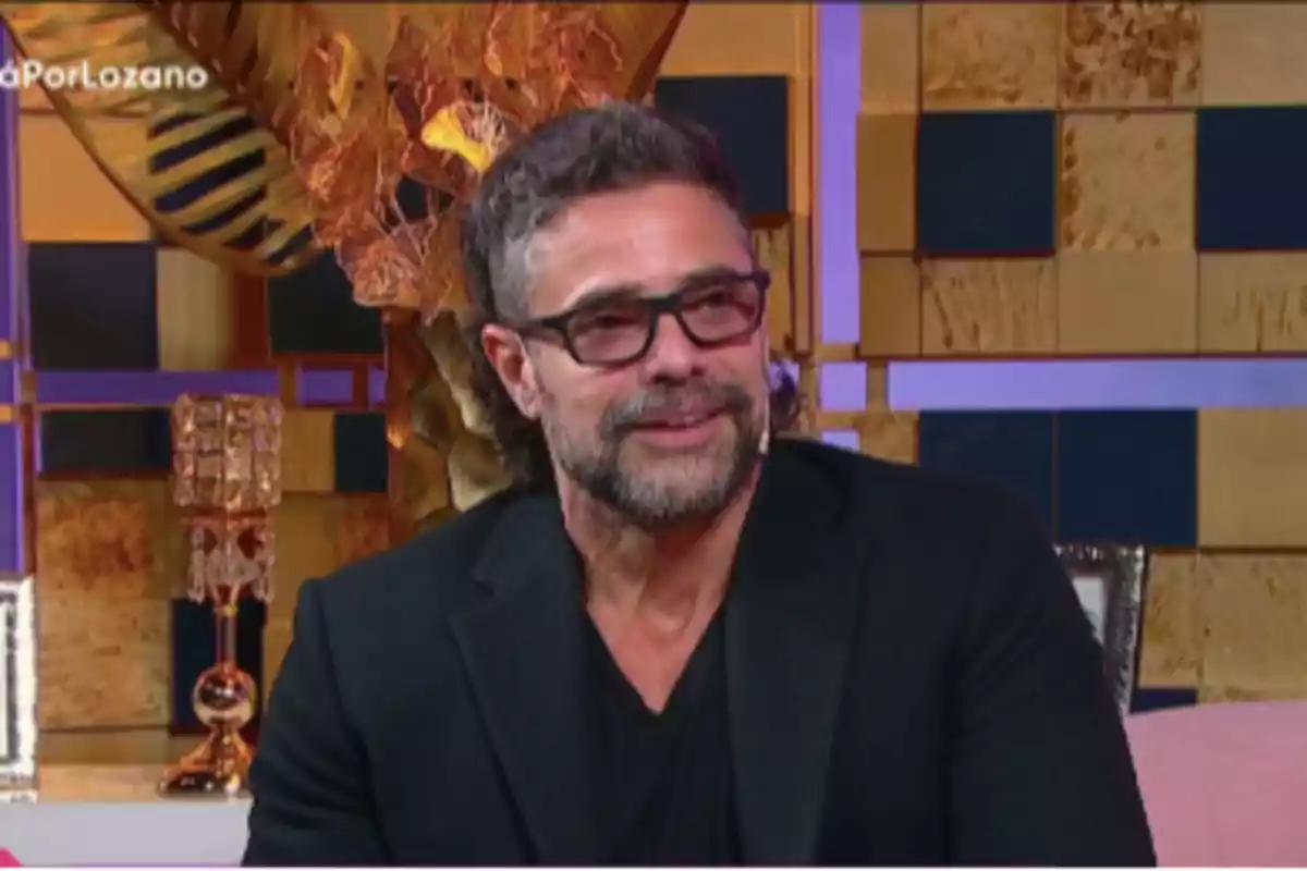 Hombre con barba y gafas oscuras sentado en un set de televisión con fondo decorativo dorado y azul