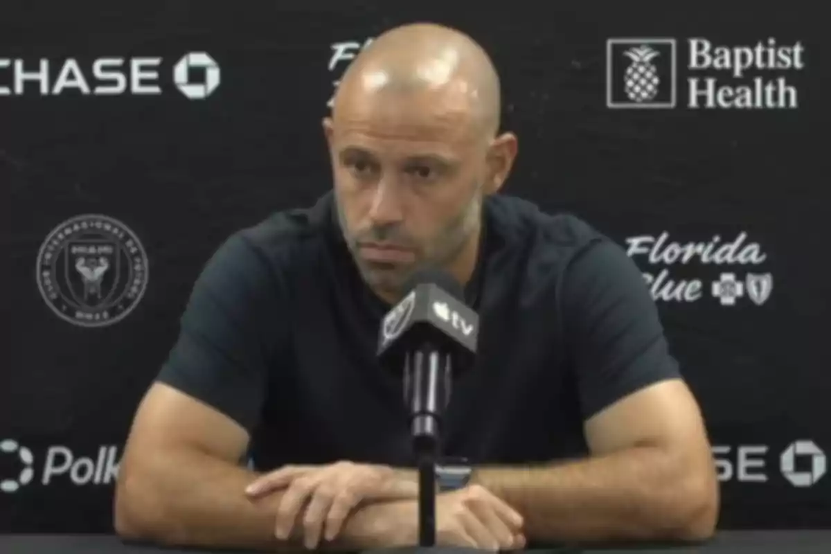 Hombre calvo con barba corta y camiseta oscura sentado frente a un micrófono durante una conferencia de prensa con fondo de logotipos de patrocinadores y equipo de fútbol Hombre calvo con barba corta y camiseta oscura sentado frente a un micrófono durante una conferencia de prensa con fondo de logotipos de patrocinadores y equipo de fútbol