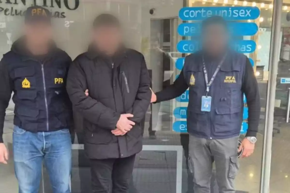 Tres personas con los rostros difuminados posan frente a una peluquería, dos de ellas llevan chalecos de la policía federal argentina y la persona del centro está vestida de negro.