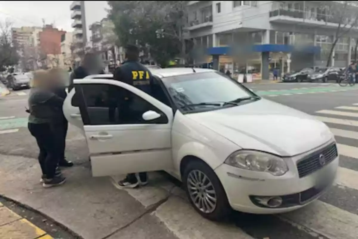 Un auto blanco detenido en una esquina con la puerta abierta y varias personas, una de ellas con chaqueta de la PFA, interactuando junto al vehículo en un entorno urbano.
