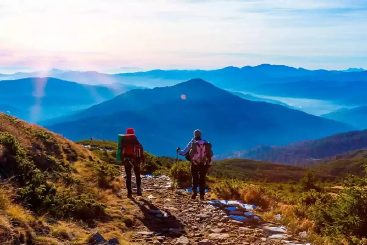 Dos personas con mochilas caminando por un sendero de montaña rodeado de vegetación con un paisaje de montañas al fondo bajo un cielo despejado