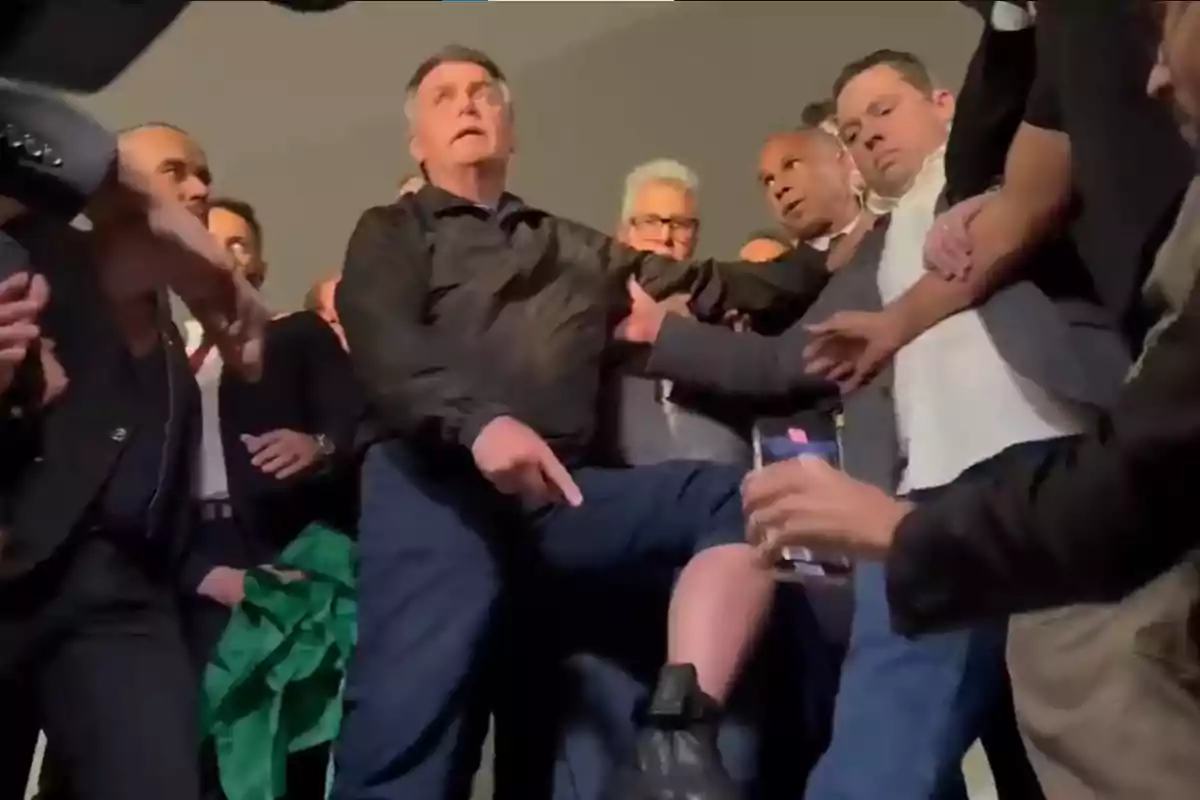 Un hombre rodeado de varias personas señala su pierna, la cual está levantada y muestra un dispositivo negro en el tobillo, mientras algunos observan y otros graban con el celular