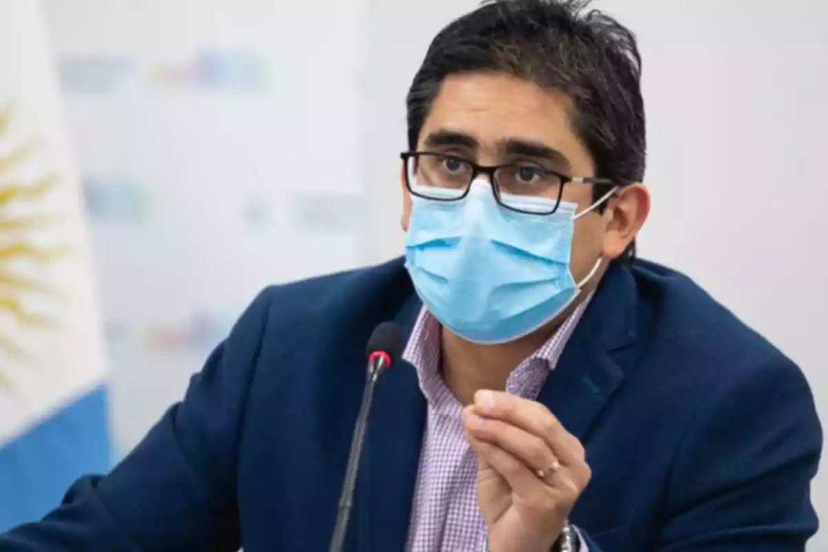 Hombre con gafas y mascarilla médica hablando frente a un micrófono en un entorno formal Hombre con gafas y mascarilla médica hablando frente a un micrófono en un entorno formal