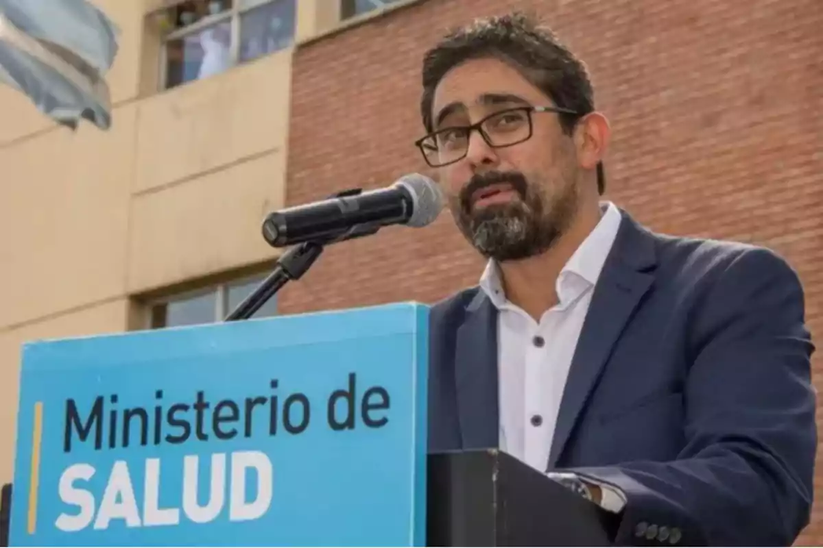 Man with a beard and glasses speaking into a microphone in front of a Ministry of Health podium with a building in the background Man with a beard and glasses speaking into a microphone in front of a Ministry of Health podium with a building in the background