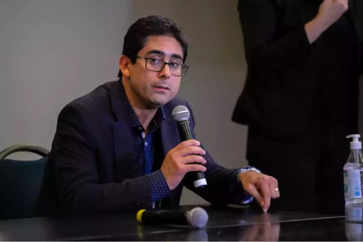 Man with glasses and a dark jacket speaking at a conference while holding a microphone, with a bottle of hand sanitizer on the table. Man with glasses and a dark jacket speaking at a conference while holding a microphone, with a bottle of hand sanitizer on the table.