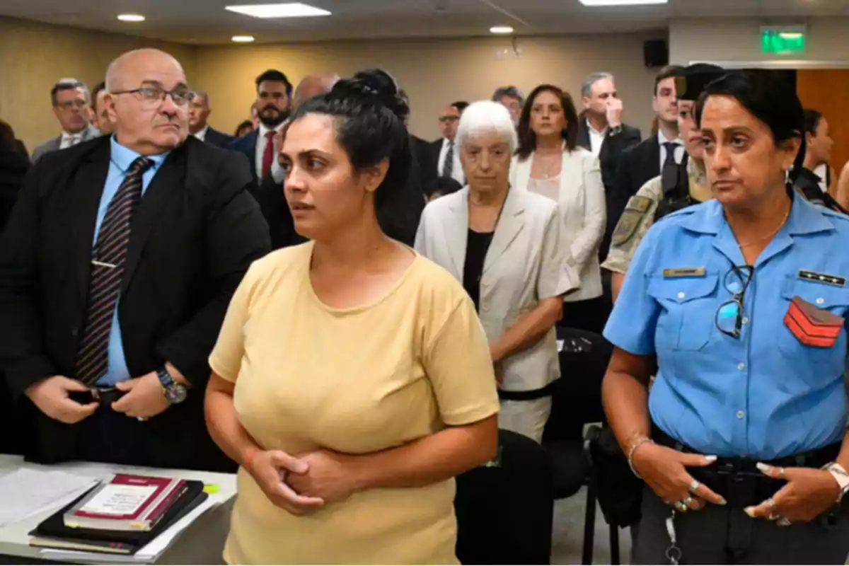 Mujer de camiseta beige de pie en una sala de audiencias rodeada de personas, incluyendo una oficial de policía y varios asistentes formales al fondo