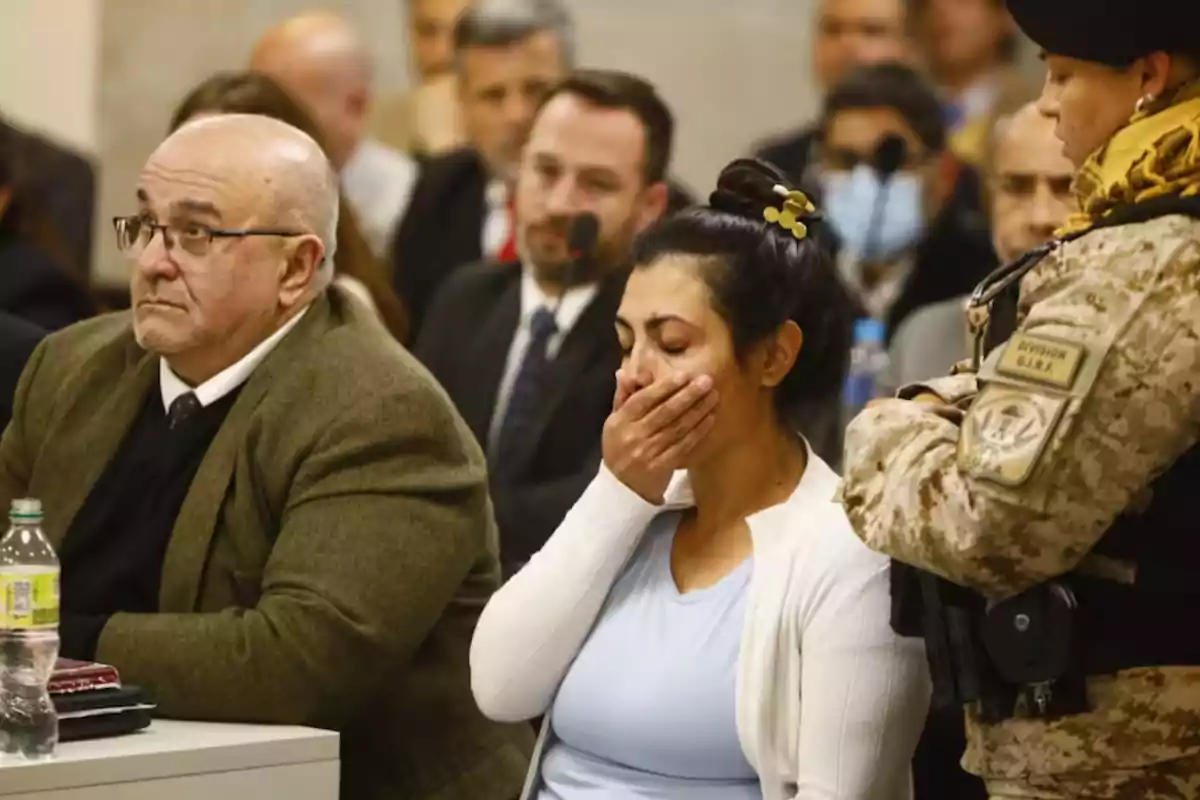 Mujer visiblemente emocionada se cubre la boca con la mano mientras está sentada junto a un hombre en una sala de audiencias rodeada de personas y una guardia uniformada Mujer visiblemente emocionada se cubre la boca con la mano mientras está sentada junto a un hombre en una sala de audiencias rodeada de personas y una guardia uniformada