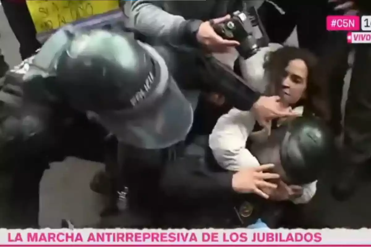 Policías forcejean con una persona durante una manifestación mientras varias manos intentan intervenir y una cámara capta el momento