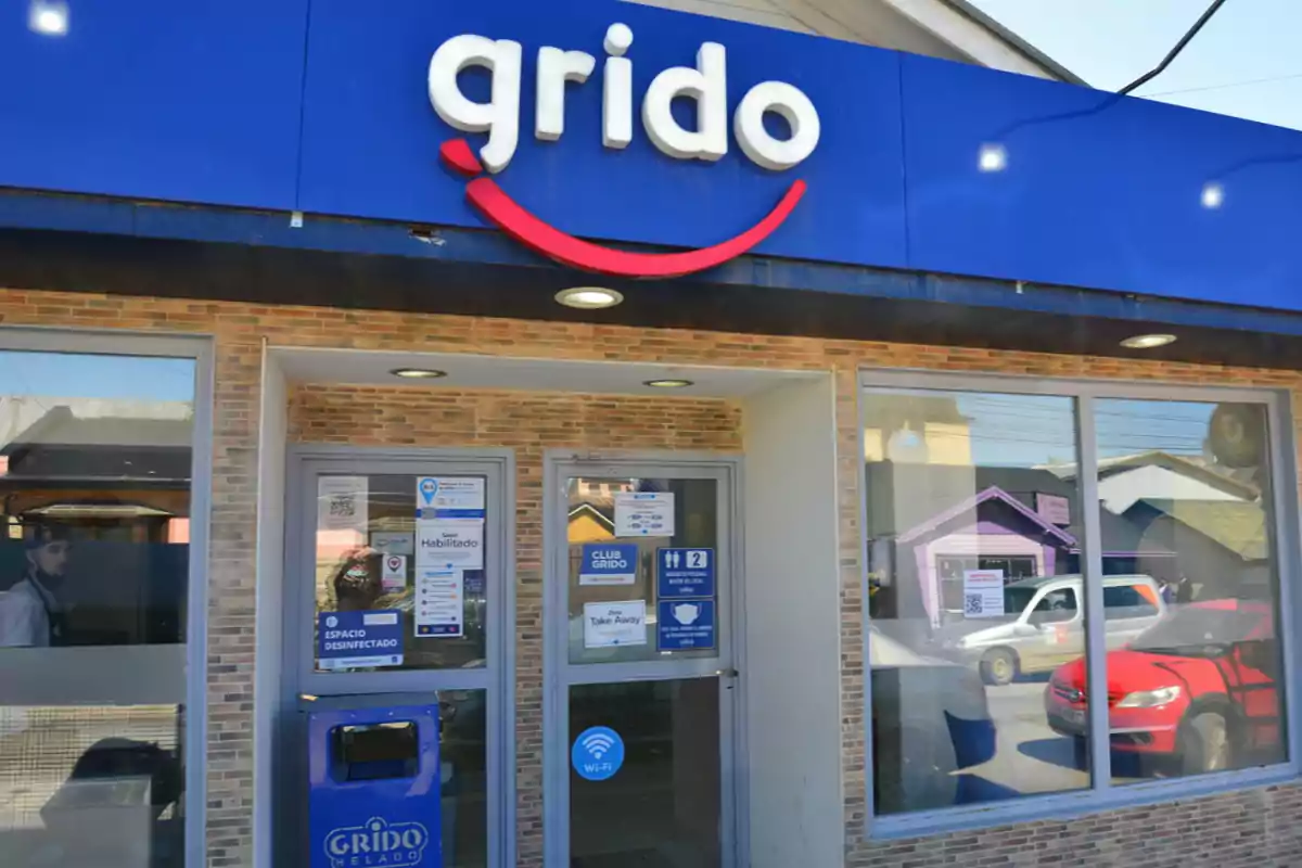 Fachada de una heladería Grido con cartel azul y letras blancas, puerta de entrada de vidrio y un tacho de basura azul en la entrada