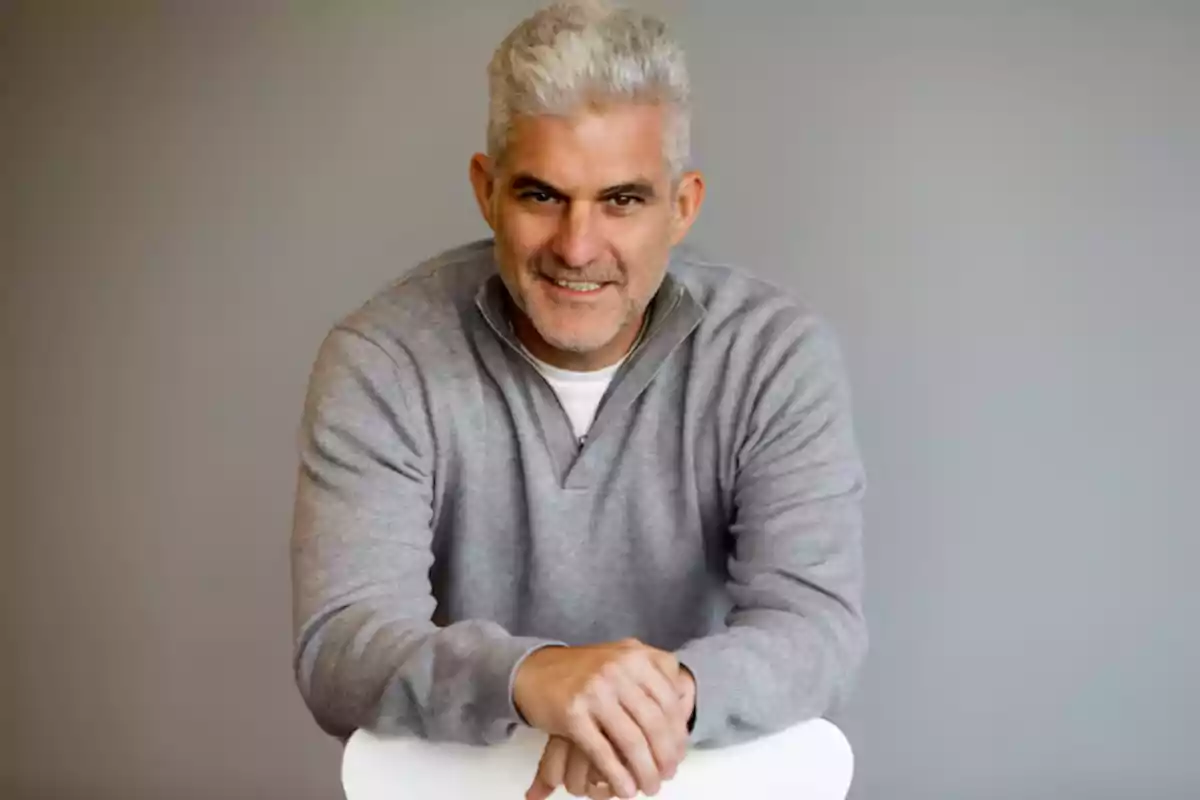 Hombre de cabello canoso y barba corta sonriendo mientras se apoya en el respaldo de una silla blanca, vistiendo un suéter gris y con fondo gris liso Hombre de cabello canoso y barba corta sonriendo mientras se apoya en el respaldo de una silla blanca, vistiendo un suéter gris y con fondo gris liso