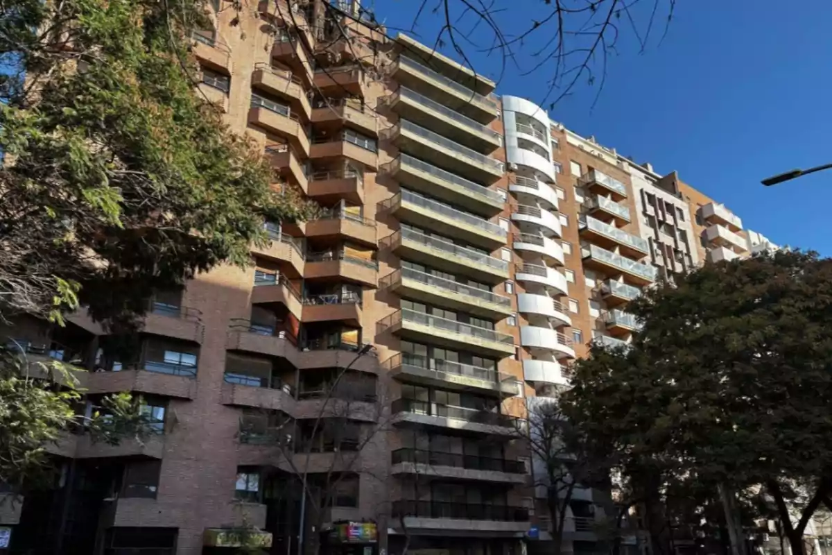 Edificio de departamentos de varios pisos con balcones y árboles en primer plano bajo un cielo despejado