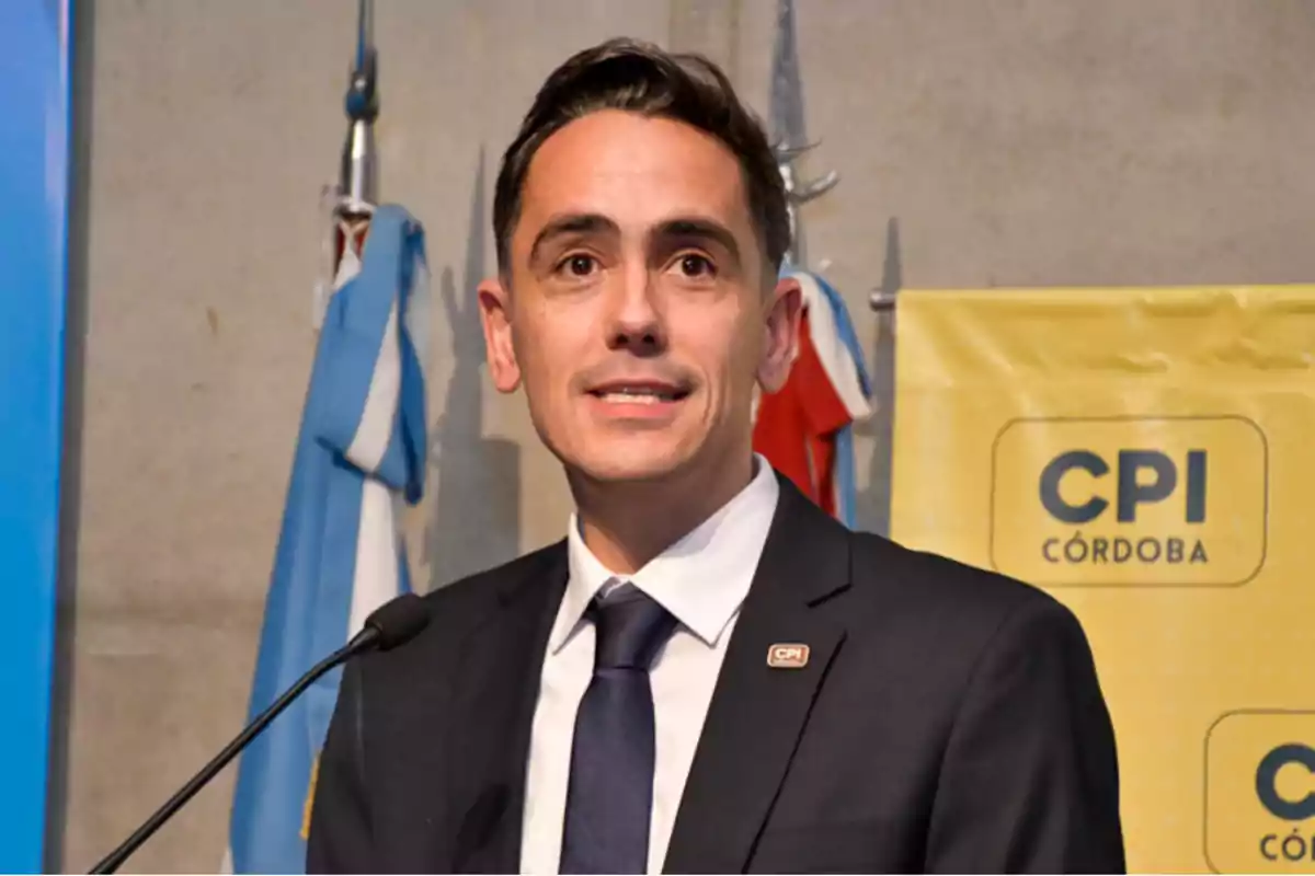 Hombre de traje hablando en un podio con banderas y un cartel amarillo de CPI Córdoba de fondo Hombre de traje hablando en un podio con banderas y un cartel amarillo de CPI Córdoba de fondo