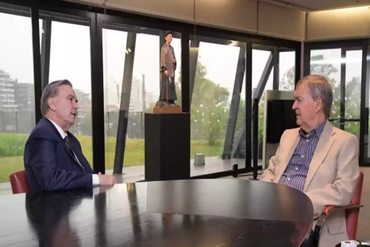 Two elderly men sit facing each other at a round table in a modern office with large windows and an outside view; in the background, there is a sculpture on a pedestal.