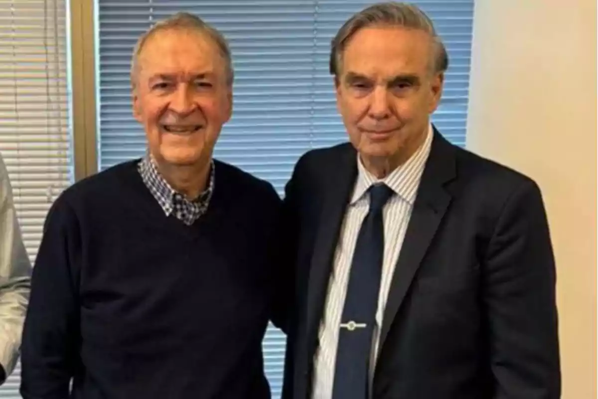 Two older men pose together in front of a window with blinds; one is wearing a dark sweater and a plaid jersey, the other is dressed in a suit and tie. Two older men pose together in front of a window with blinds; one is wearing a dark sweater and a plaid jersey, the other is dressed in a suit and tie.