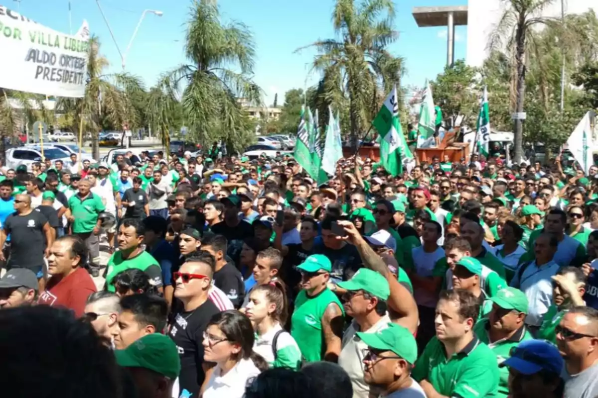 Multitud de personas reunidas al aire libre durante una manifestación, muchas de ellas vestidas con camisetas y gorras verdes, algunas sosteniendo banderas y pancartas, con palmeras y autos al fondo.