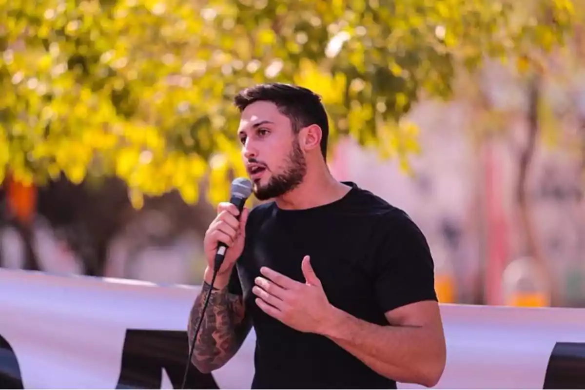 Hombre joven con barba y camiseta negra hablando por micrófono al aire libre frente a un fondo de árboles desenfocados Hombre joven con barba y camiseta negra hablando por micrófono al aire libre frente a un fondo de árboles desenfocados