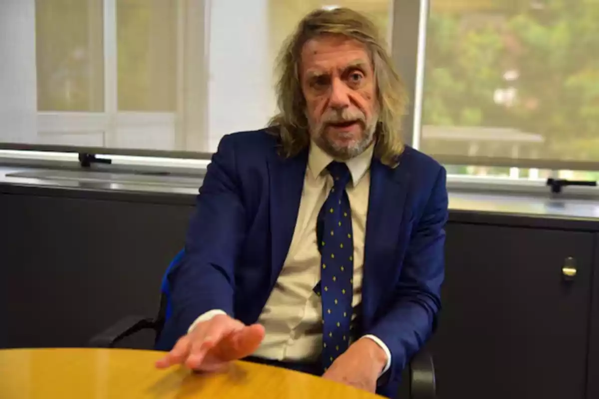 Hombre de cabello largo y canoso con barba, vestido con traje azul y corbata, sentado en una oficina mientras extiende una mano sobre una mesa Hombre de cabello largo y canoso con barba, vestido con traje azul y corbata, sentado en una oficina mientras extiende una mano sobre una mesa